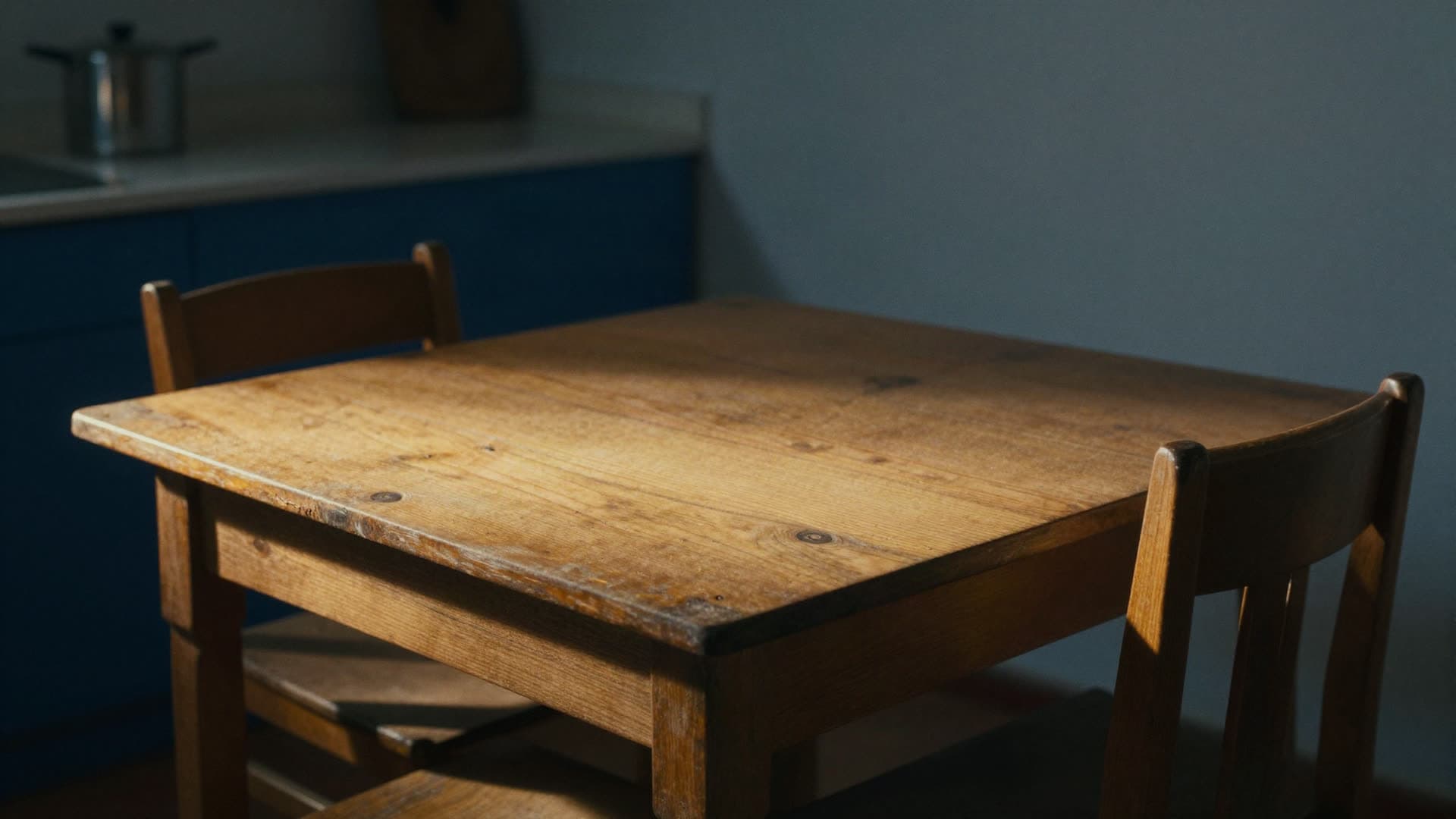 A simple worn kitchen table with two empty chairs, late afternoon light falling across the wood.
